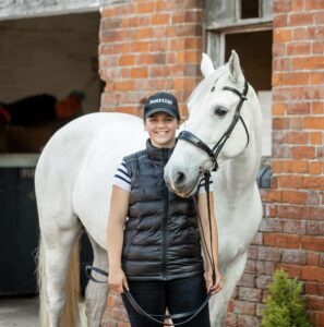 Photo of Amy stood looking at camera with Grey horse wearing a bridle