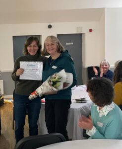 Two women stood together holding a certificate and bunch of flowers smiling at the camera
