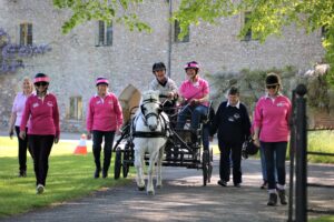 Grey pony pulling a four wheeled carriage with coach and participant onboard. Surrounded by a team of 5 supporters on foot wearing pink tops. 