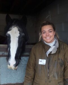 Rachel wearing a white polo neck and brown waxed jacket, smiling to camera standing outside a stable, with a black pony with a white blaze looking over the door.