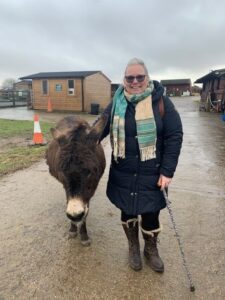 Mel looking to camera, standing outside next to a small brown donkey. Mel wears glasses, a stripey scarf and a long blue puffer coat.